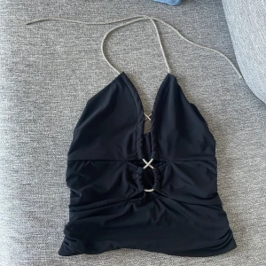 Svart halterneck topp med strassdetaljer - Supersnygg svart halterneck topp med glittriga strassband som knyts i nacken. Djup v-ringning och två dekorativa strassringar framtill ger en extra festlig vibe. Perfekt för dig som vill sticka ut och glänsa lite extra.