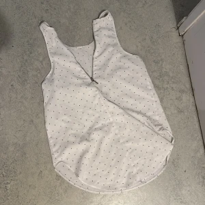 Vit prickig linnetopp med dragkedja - Säljer en vit linnetopp med små svarta prickar och en snygg dragkedja framtill. Toppen är ärmlös med breda axelband och har en rundad nederkant. Perfekt för en fräsch och enkel look under varma dagar.