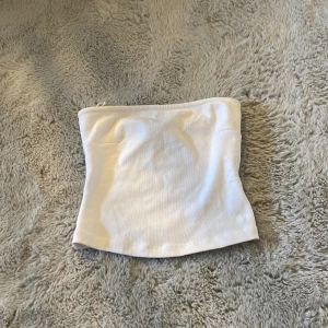 Tubtopp - Säljer en enkel och stilren vit  tubtopp från Gina Tricot. Toppen är ärmlös och axelbandslös, gjord i mjuk bomull med en lätt ribbad struktur. Perfekt för att skapa en clean och fräsch look till sommaren. I strl 146/152. Kontakta mig vid frågor eller fler bilder, alldrig använd.