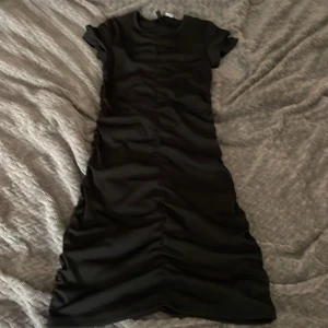 Svart långklänning  - Svart långklänning från Divided med korta ärmar och snygg rynkad detalj längs hela framsidan. Klänningen har en figurnära passform och är tillverkad i ett stretchigt material som sitter bekvämt. Perfekt för dig som gillar en stilren och modern look.