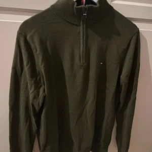 Olivgrön half zip tröja Tommy Hilfiger - Olivgrön långärmad tröja från Tommy Hilfiger med half zip och hög krage. Liten broderad logga på bröstet och snygga ribbade detaljer vid krage och ärmslut. Tillverkad i mjuk bomull, perfekt för lager-på-lager under höst och vinter.