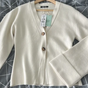 Vit kofta med knappar från Gina Tricot - Vit kofta från Gina Tricot med v-ringning och bruna knappar framtill. Koftan har långa ärmar med breda muddar och är tillverkad av mjuk stickad polyesterblandning, varav 51% är återvunnen polyester. Perfekt för lager-på-lager och enkel att matcha.