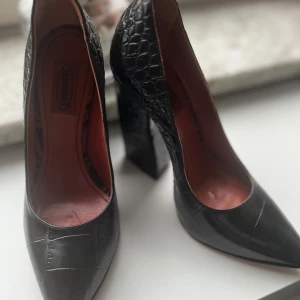 Svarta pumps i skinn från Missoni - Chunky svarta pumps från Missoni i präglat skinn med croco-mönster. Skorna har spetsig tå och hög, bred klack som ger en cool statement-look. Insidan är rödbrun och materialet känns lyxigt. Perfekta för dig som vill sticka ut med elegans.