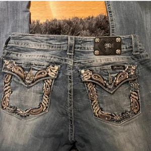 Miss Me jeans med broderade fickor - Snygga blå Miss Me jeans med låg midja och bootcut. Jeansen har detaljerade broderier och paljetter på bakfickorna samt slitningar för en cool look. Klassisk femficksmodell med silverfärgade knappar och logga bak. Perfekta för dig som gillar statement-jeans.