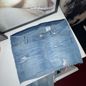 Blå jeanskjol med slitningar - Snygg blå jeanskjol från Addison med rå kant nertill och slitna detaljer framtill. Klassisk femficksmodell med bälteshällor och knappstängning. Perfekt för dig som gillar en avslappnad och trendig stil.
