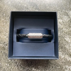 Svart flätat armband Tommy Hilfiger - Stilrent svart armband från Tommy Hilfiger med flätad design i skinn och ett silverfärgat magnetlås där Hilfiger-loggan är ingraverad. Perfekt accessoar för en clean och modern look. Levereras i originalask. Aldrig använd