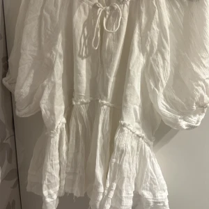 Vit bohemisk blus - Superfin vit blus i luftig bomull med volanger och knytning vid halsen. Den har vida ärmar och en avslappnad passform, perfekt för varma dagar.