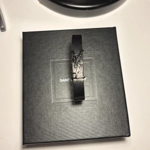 Svart Saint Laurent läderarmband - Stilrent svart armband från Saint Laurent i äkta läder med ikoniska YSL-loggan i svart metall framtill. Smal och minimalistisk design som ger en lyxig känsla. Kommer i original ask. Perfekt accessoar för att lyfta din outfit. Hör av er vid frågor! Pris ej hugget i sten! Kvitto finns så bara komma privat!