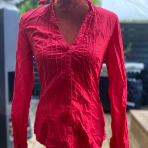 Röd skjorta från edc, storlek M - Röd skjorta med långa ärmar och v-ringning. Skjortan har knappar framtill och detaljerade veck på framsidan. L: 67 cm.  ﻿Bystvidd: 50 cm ﻿100% bomull  Inga bilder när den sitter på mig! Katter finns i hemmet!