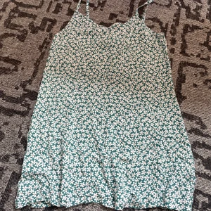 Grön blommig sommarklänning Sinsay - Supersöt grön kortklänning från Sinsay med vita blommor, smala axelband och snörning framtill. Perfekt för varma dagar och har en lätt och luftig känsla. Klänningen har v-ringning och är ärmlös, vilket gör den extra somrig och chill.