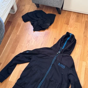En Svart tröja från Hugo Boss - Svart tröja från Hugo Boss med blå detaljer och dragkedja. Jackan har broderad logga på bröstet, huva med snörning och blått innerfoder. Axelpartiet är i ett diskret rutigt tyg och ärmarna har blåa detaljer. Perfekt för en chill och stilren look.