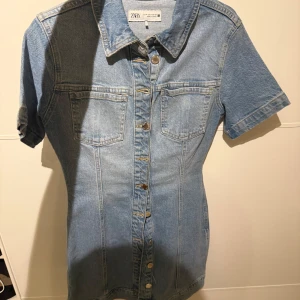 Jeansklänning från Zara med knappar - Snygg ljusblå jeansklänning från Zara med kort ärm och klassisk skjortkrage. Klänningen har knappar hela vägen framtill och två bröstfickor. Perfekt för en chill och trendig look.