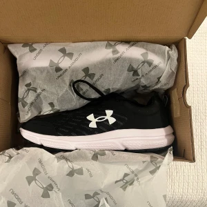 Svarta sneakers från Under Armour - Snygga svarta sneakers från Under Armour med vit sula och stor vit logga på sidan. Ovandelen är i mesh för bra andningsförmåga och skon har snörning. Perfekta för träning eller en sportig vardagslook. Sulan är designad för bra grepp och komfort. (Helt oanvända)