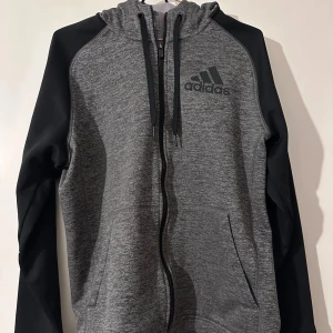 Grå hoodie från Adidas - Säljer en snygg och bekväm grå hoodie från Adidas med svarta ärmar och dragkedja. Perfekt för kyliga dagar med sitt mjuka och varma material. Adidas-loggan pryder bröstet och den har en praktisk huva med dragsko. Passar både till vardags och träning!