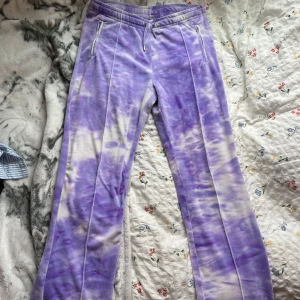 Lila tie-dye Juicy Couture mjukisbyxor - Supermjuka lila och vita tie-dye mjukisbyxor från Juicy Couture med glittrande 'Juicy' i strass på baksidan. Byxorna har dragsko i midjan, raka ben och två fickor med dragkedja framtill. Perfekta för en chill och trendig look. Lång ben.