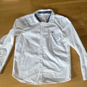 Vit skjorta från H&M i bomull - Klassisk vit skjorta från H&M i storlek 146. Skjortan har lång ärm, knappar framtill och en liten broderad detalj på bröstet. Tillverkad i mjuk bomull och har en stilren krage med kontrastfärg på insidan. Använd mycket men utan hål och ren.                                    Axelbredd: 35 cm.                                  Ärmlängd: 53 cm.                                             Säljer för att den är för liten. Kontakta gärna vid intresse! 