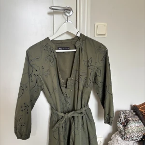 Olivgrön kortklänning från Zara - Olivgrön kortklänning från Zara med broderade detaljer och hålmönster. Klänningen har långa ärmar, knappar framtill och ett matchande knytbälte i midjan. Perfekt för dig som gillar en avslappnad men ändå snygg stil.