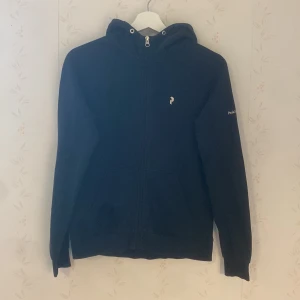 Svart hoodie från Peak Performance junior - Svart hoodie från Peak Performance med dragkedja och huva. Klassisk logga på bröstet och ärmen. Mjuk insida och känguruficka framtill. Perfekt för chill eller när du vill ha en enkel, stilren look. Junior storlek M cirka 155-165 cm