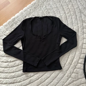 Svart långärmad topp med v-ringning - En svart långärmad topp från Gina Tricot med snygg v-ringad halsringning. Modellen är figurnära och har en enkel, stilren design som passar perfekt till jeans eller kjol. Mjuk och stretchig i materialet för en bekväm känsla.