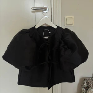 Svart bolerotopp från Gina Tricot - Supersnygg svart bolerotopp från Gina Tricot med puffiga korta ärmar och två knytband framtill. Toppens material har en lätt glansig finish som ger en lyxig känsla. Perfekt för dig som vill sticka ut med en trendig och cool look.