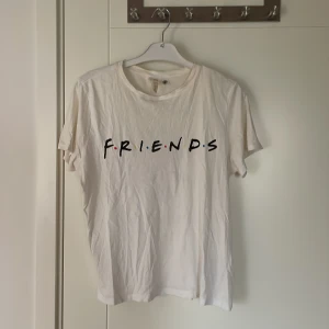 Vit Friends t-shirt från H&M - Vit t-shirt från H&M med Friends-tryck på bröstet och text på ryggen inspirerad av karaktärerna från serien. Klassisk passform, rund hals och korta ärmar. Tillverkad i mjuk bomull, perfekt för dig som älskar 90-talsvibbar och tv-serier.