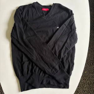 Svart stickad tröja J.Lindeberg M - Svart stickad tröja från J.Lindeberg med v-ringning och diskret JL-logo på bröstet. Klassisk modell med ribbade muddar och lång ärm, perfekt för lager-på-lager. Snygg och enkel att matcha till det mesta.