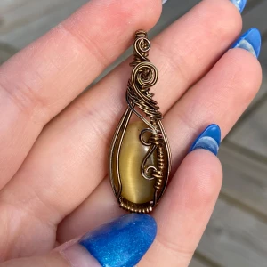 Wirewrapat berlock med gul sten - Unikt handgjort halsband med en droppformad gul sten infattad i konstnärligt snirklig wirewrap av bronsfärgad metall. Stenen har en mjuk glans och metalltråden är virad i spiraler och böjda former som ger ett boho-vibe. Perfekt för dig som gillar statement-smycken.