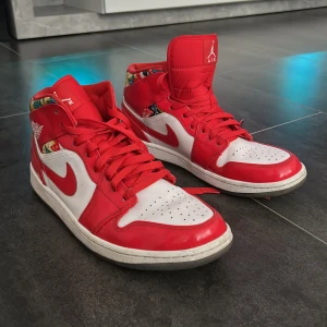 Nike Air Jordan 1 röda och vita - Säljer ett par Nike Air Jordan 1 med röd och vit färg, klassisk swoosh och coolt mönster i färgglada zigzag-detaljer runt ankeln. Skorna har snörning, rund tå och hög siluett. Perfekta för dig som vill sticka ut med en ikonisk sneaker.