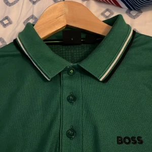 Grön pikétröja från BOSS - Snygg grön pikétröja med fräscha vita detaljer från BOSS med klassisk krage och knäppning framtill. Tröjan har svarta och vita ränder på krage och ärmslut samt diskret BOSS-logga på bröstet och vid nacken Perfekt för en clean och sportig look, med storlek S passar Xs också men sitter perfekt och passar  bäst på S, använd 3 gånger ser helt ny ut, en ny ligger på 1250 men säljer den för 500.