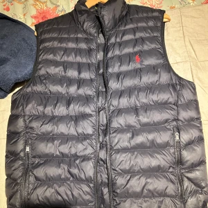Svart dunväst från Ralph Lauren - Snygg svart dunväst från Ralph Lauren med horisontella quiltade sömmar och klassisk röd logga på bröstet. Västen har hög krage, två fickor med dragkedja och stängs med dragkedja framtill. Perfekt att slänga över en hoodie för extra värme.