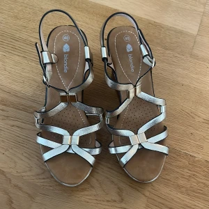 Chrome kilklackar från Blondie - Snygga kilklack från Blondie i metallic silver. Remmar i skimrande syntet med ormskinnsstruktur, öppen tå och justerbart spänne vid ankeln så även användbara vid strl 39. Perfekta för lit sommarkvällar.🪩