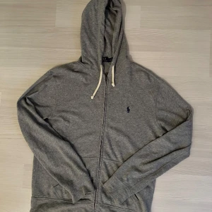 Grå hoodie från Polo Ralph Lauren - Grå hoodie från Polo Ralph Lauren med dragkedja och vita snören, riktigt fet hoddie som tyvärr inte används så ofta, Strolek S, Pris 300 kr 