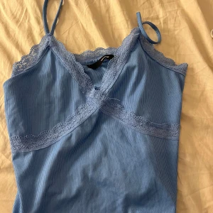 Blå ribbad linnetopp från Vero Moda - Supersöt ljusblå ribbad linnetopp från Vero Moda med spetsdetaljer längs urringningen och under bysten. Smala axelband och omlottkänsla framtill gör den extra fin. Perfekt att styla med jeans eller kjol för en chill look.