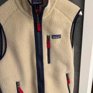 Beige fleeceväst från Patagonia - Säljer en snygg beige fleeceväst från Patagonia med marinblå detaljer och dragkedjor. Västen har flera praktiska fickor med röda dragkedjor och en hög krage för extra värme. Perfekt för lager-på-lager under kyliga dagar. Nypris- 1400kr