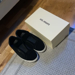 Svarta sneakers från Axel Arigato - Stilrena svarta sneakers från Axel Arigato med vit platt sula. Skorna har en minimalistisk design utan snörning, med ovandel i mocka och detaljer i skinn. Perfekta för dig som gillar en clean och modern look.