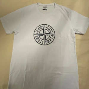 Vit Stone Island t-shirt med tryck - Helt ny och oanvänd vit t-shirt från Stone Island med stort svart logotryck på bröstet. Klassisk rund halsringning och korta ärmar. Tillverkad i 100% bomull för en skön och luftig känsla. Perfekt för dig som gillar streetwear och vill sticka ut med en ikonisk look. Liten i storlek och passar M.