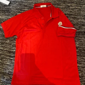 Röd Moncler pikétröja - Snygg röd pikétröja från Moncler med klassisk krage, knäppning framtill och broderad logga på ärmen. Tröjan har vita och svarta detaljer på ärmslutet och är tillverkad i mjuk bomull. Perfekt för dig som vill ha en stilren och sportig look. Storlek M