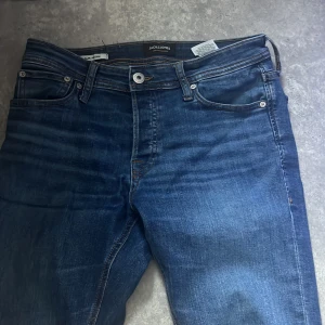 Blå jeans från Jack & Jones, 30/34 - Säljer ett par klassiska blå jeans från Jack & Jones i modellen Glenn. Jeansen har fem fickor, snygga kontrastsömmar och en smal passform. Tillverkade i bomull med lite stretch för extra komfort. Perfekta till sneakers eller boots.