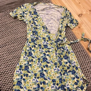 Blommig omlottklänning Pull&Bear - Superfin kort omlottklänning från Pull&Bear med blommigt mönster i blått, gult och vitt. Klänningen har korta ärmar, knytband i midjan och v-ringning. Perfekt för sommarens alla tillfällen och ger en fräsch och färgglad look.