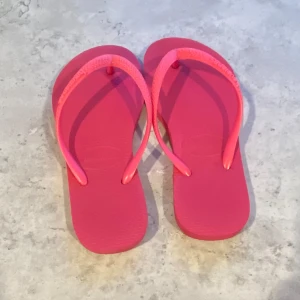 Rosa flip-flops från Havaianas - Snygga och klassiska flip-flops från Havaianas i en stark rosa färg. De har en enkel design med mjuka remmar och platt sula, perfekta för stranden eller poolen. Tillverkade i syntetmaterial som tål vatten och är lätta att bära. 