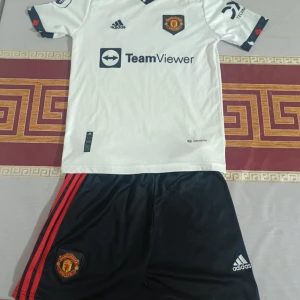 Manchester United bortatröja Adidas - Manchester Uniteds bortaställ från Adidas. Tröjan är vit med svarta och röda detaljer, kortärmad och har klubbmärke samt TeamViewer-tryck på bröstet. Matchande svarta shorts med röda ränder och klubbmärke.