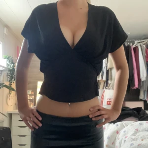 Zara topp - Svart croppad omlottopp med djup v-ringning och korta ärmar. Toppen har en figurnära passform och smockad detalj i ryggen som ger en snygg siluett. Perfekt att styla med höga byxor eller kjol för en trendig look.