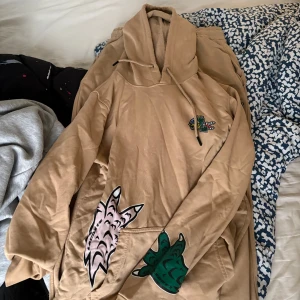 Beige hoodie från Lacoste med tryck - Säljer en beige hoodie från Lacoste med coola tryck av gröna och rosa krokodilklor på framsidan. Tröjan har huva med snörning och en broderad Lacoste-logga på bröstet. Perfekt för dig som gillar streetwear och vill sticka ut lite extra.