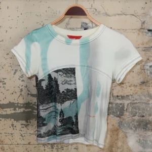 Ekhaus Latta t-shirt med landskapsprint - Nypris 2000kr. Unik t-shirt från Ekhaus Latta med coolt landskapsprint i svart och turkos på vit botten. Toppen har korta ärmar, rund hals och kontrastsömmar. Materialet är mjuk bomull och passformen är normal. Perfekt för dig som gillar konstnärliga och annorlunda plagg.