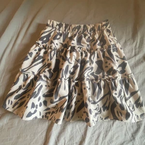 Beige volangkjol med leopardmönster - Superfin kort volangkjol i beige med svart leopardmönster. Kjolen har flera lager med volanger och resår i midjan för en bekväm passform. Perfekt för dig som vill sticka ut med en trendig look.