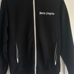 Svart Palm Angels track jacket M - Svart track jacket från Palm Angels med vita ränder längs ärmarna och vit dragkedja framtill. Jackan har två fickor med dragkedja och logga på bröstet. Använt sparsamt! Köpt från Farfecht.