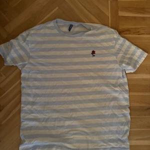 Randig t-shirt med broderad ros - Vit och ljusgrå randig t-shirt från Cubus med korta ärmar och en liten broderad röd ros på bröstet. Skön passform och klassisk rund halsringning. Perfekt för dig som gillar stilrena detaljer och en chill vibe.