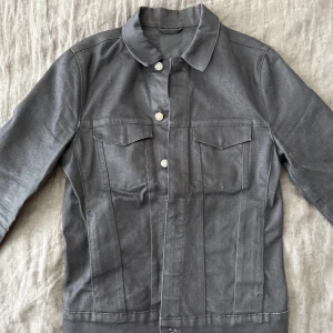 Svart denimjacka från J.Lindeberg - Snygg svart denimjacka från J.Lindeberg med klassisk krage, silverfärgade knappar och två bröstfickor med lock. Jackan har raka linjer och en stilren look som passar perfekt till streetwear eller casual outfits.