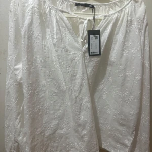 Vit broderad blus från LAGER157 - Superfin vit blus från LAGER med broderade blommor över hela tyget. Långärmad modell med v-ringning och knyt fram. Luftig och lätt i bomull, perfekt för vår och sommar. Söt och romantisk vibe som passar till jeans eller kjol.