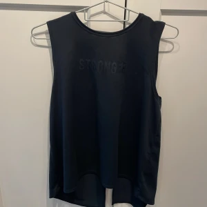 Svart linnetopp från Stronger - Snygg svart linnetopp från Stronger med diskret logga framtill. Modellen är ärmlös med rund halsringning och längre baktill med en cool slits i ryggen. Perfekt för träning eller när du vill ha en sportig look.